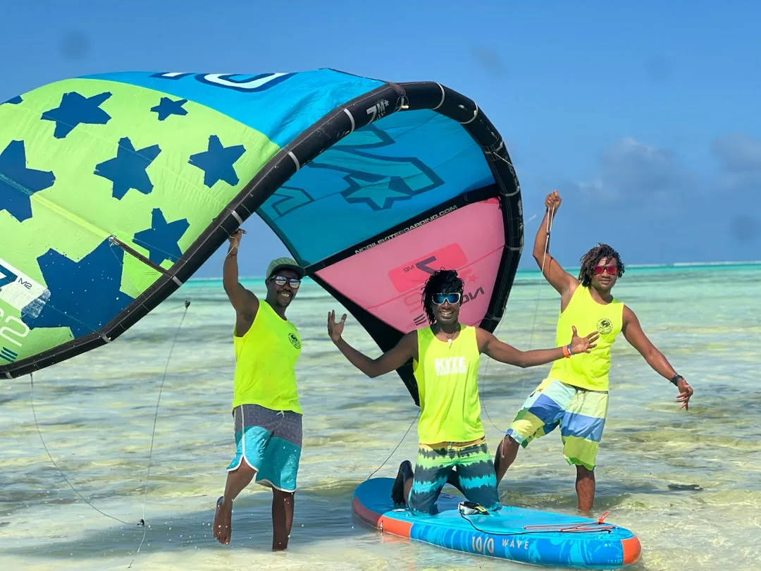 Three Mawimbi teachers in yellow vests hold up a kite in the shallow turquoise lagoon, a board at their feet