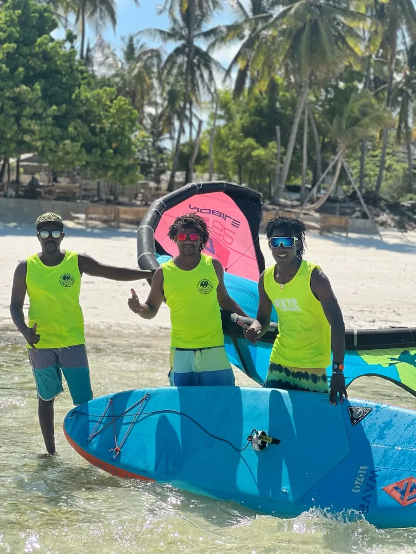 The Mawimbi teachers in yellow vests on the beach under palm trees, with a kite and a board